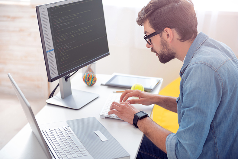homem branco, de óculos, digitando no computador. À esquerda dele, um notebook. À direita, um tablete. O homem veste uma camisa jeans e usa um relógio no pulso esquerdo. O filtro web garante mais produtividade para a sua empresa. 