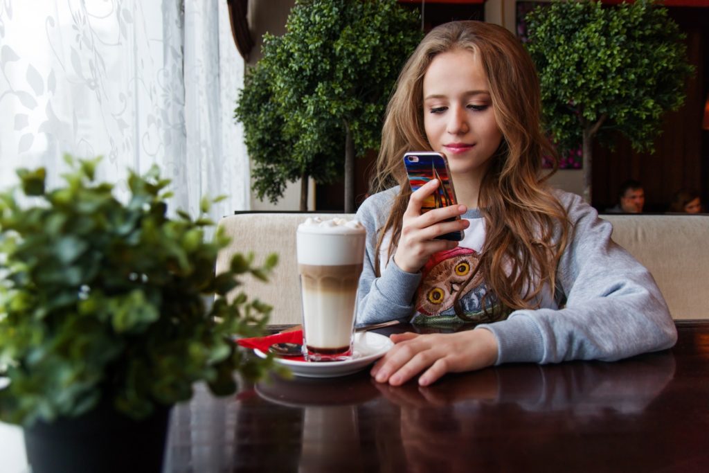 Era mobile: menina mexendo no celular sentada à mesa, com um copo de café em frente.