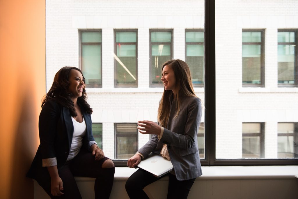 Melhorar a experiência do cliente: duas mulheres conversando, enquanto estão sentadas no batente de uma janela. As duas seguram um copo de café.