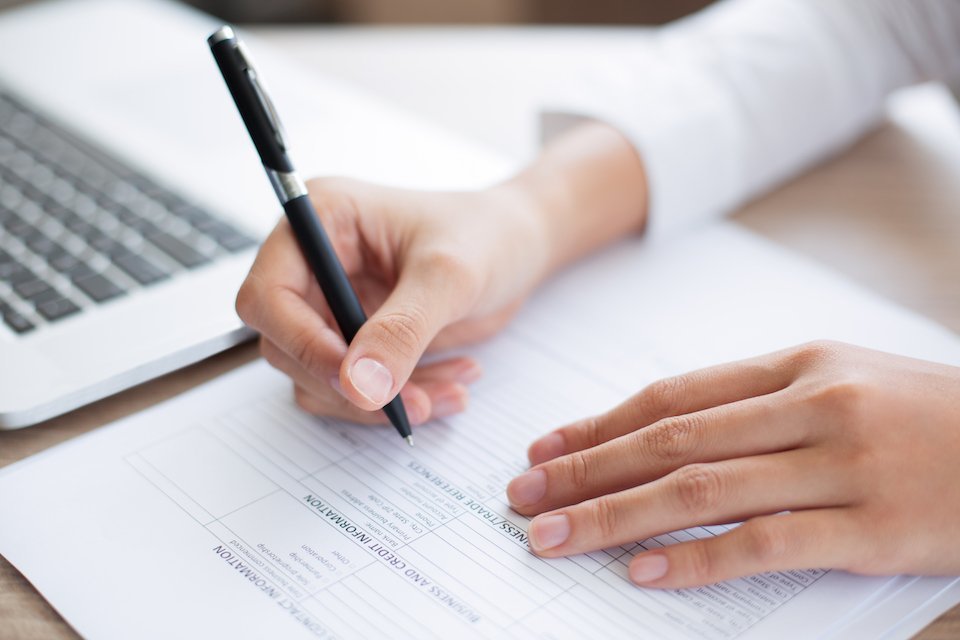 Uma pessoa preenche um formulário que encontra-se em cima de uma mesa de madeira, ao lado de um notebook. Na mão direita, uma caneta enquanto a mão esquerda repousa sobre o papel.