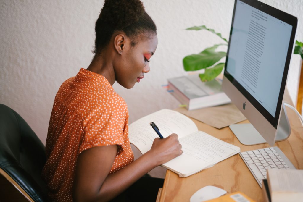 mulher em frente ao computador, fazendo uma anotação no bloco de notas.