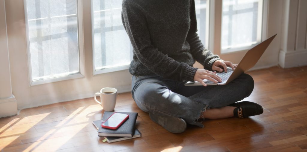 Imagem de uma pessoa sentada no chão com o computador no colo e uma xícara de café ao lado. 