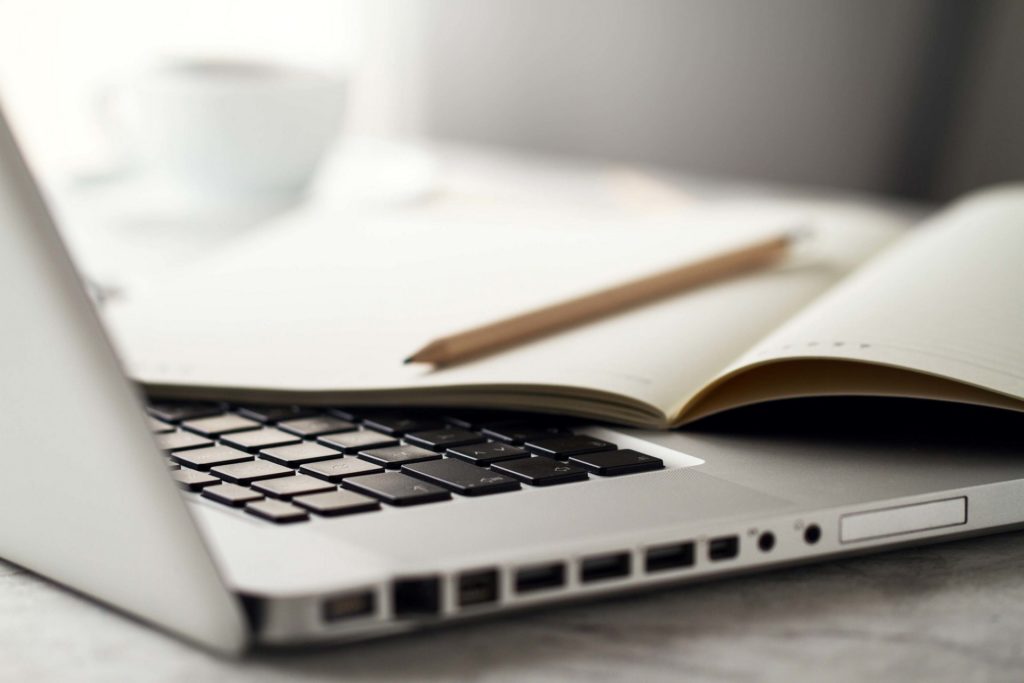 Imagem de um notebook aberto e um caderno e um lápis em cima do teclado. 