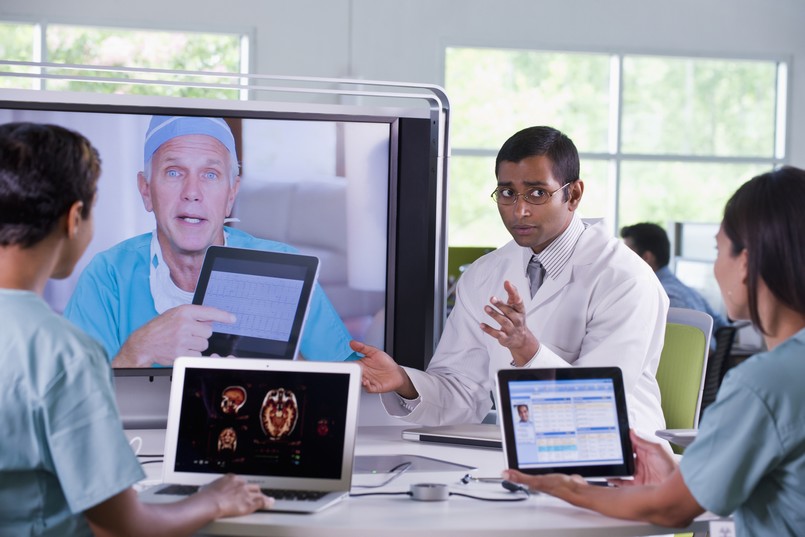 Imagem de um médico e dois enfermeiros sentados com tablets nas mãos enquanto olham para um médico cirurgião que aparece em uma televisão em videoconferência para simbolizar a tecnologia na enfermagem. 