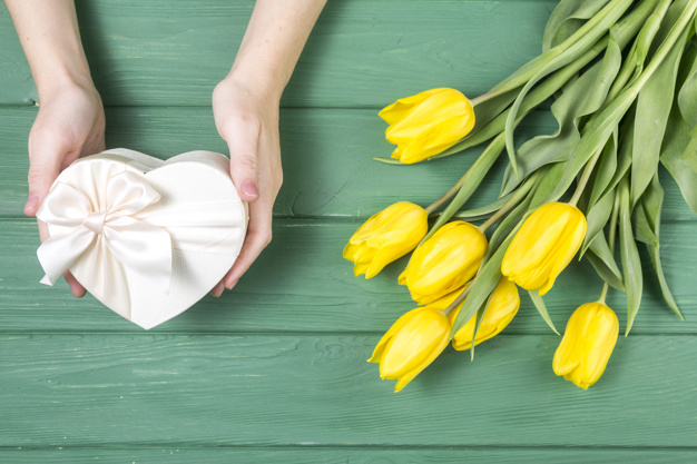 Imagem de duas mãos segurando uma caixa de presente em formato de coração e flores amarelas ao lado para simbolizar o dia dos namorados. 