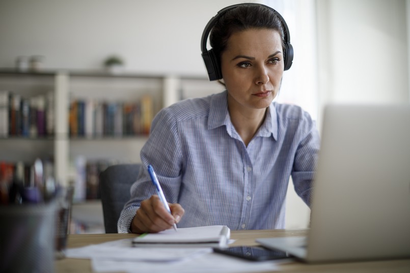 Imagem de uma mulher com fones de ouvido olhando um computador e escrevendo em um caderno. 