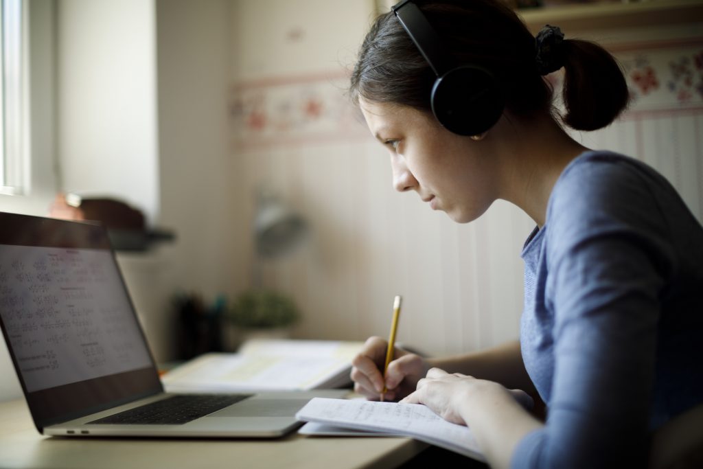 Mulher de fone olhando para o notebook e fazendo anotações enquanto realiza o curso online